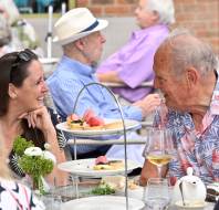 Two people sit at a table outdoors, smiling and talking, with plates of desserts and glasses of wine in front of them. Other diners are visible in the background.