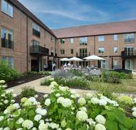 Modern brick apartment building with balconies, large windows, and a garden courtyard where people are seated under white umbrellas.
