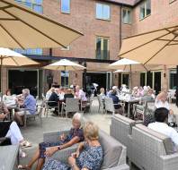 A group of people sit at tables and lounge chairs under large umbrellas on a patio outside a brick building, socializing and dining.