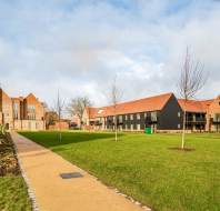 A modern residential complex with red-tiled roofs, manicured lawns, young trees, and a paved walkway under a partly cloudy sky.