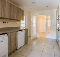 Modern galley kitchen with wood cabinets, beige countertops, built-in appliances, tiled floor, and double doors leading to a bright hallway.