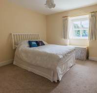 A simple bedroom with a double bed, white bedding, two pillows, a window with cream curtains, a wicker basket, and beige carpeted floor.