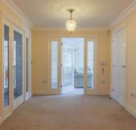 Empty beige-carpeted room with cream walls, glass double doors leading to a sunlit room, closed doors on both sides, and a ceiling light fixture.