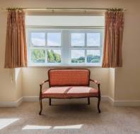 A vintage upholstered bench sits in front of a window with open curtains, letting in natural light in a neutral-colored room with carpeted flooring.