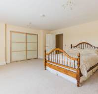 A spacious bedroom with beige walls, a wooden bed with cream bedding, sliding door closet, and light carpet flooring.