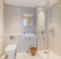 Modern bathroom with beige tiles, wall-mounted toilet, small sink with mirror, glass shower enclosure, towel rack, and a wicker waste basket on the floor.