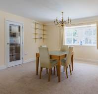 A dining room with a wooden table, four beige chairs, a gold chandelier, carpeted floor, wall shelves, and a large window with curtains letting in natural light.