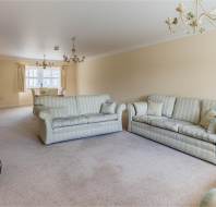 Spacious living room with beige walls, carpeted floor, two striped sofas, chandeliers, a small stove, and a dining table by the window in the background.