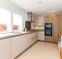 Modern kitchen with beige cabinets, built-in oven, stove, and sink under two windows; wooden accents and neutral tile flooring are present, with a small table and stools on the right.