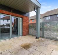 A small patio with beige paving slabs, a potted plant, and glass doors leading inside; surrounded by a fence and brick exterior walls of modern residential buildings.