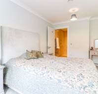 Bedroom with a large bed, two pillows, a nightstand with a radio, a dressing table with a mirror, and an open door leading to a bathroom.