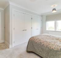 Bedroom with double bed, patterned bedspread, double wardrobe, window with Roman shade, and neutral decor. Doorway leads to dining area with table and chairs.