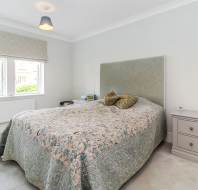 Bedroom with a double bed, patterned bedspread, two pillows, bedside table with lamp, another bedside unit with alarm clock, window with valance, and light fixture on the ceiling.