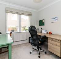 A home office with two desks, an office chair, a window with blinds, wall calendar, files, desk lamp, and various office supplies.