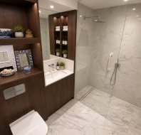 Modern bathroom with marble walls, walk-in shower, glass partition, wooden shelving, toilet, sink with mirror, and decorative items. Bright lighting and organized toiletries are visible.