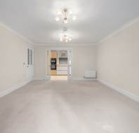 Empty room with light-colored walls and carpet, leading to a kitchen through double sliding doors.