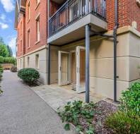 A brick apartment building with a ground-floor terrace, open glass double doors, and an adjacent stone pathway.