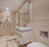 A bathroom featuring a glass-enclosed shower, a wall-mounted vanity with a sink and medicine cabinet, and a toilet flush plate on the tiled wall.