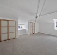 An empty attic room with white walls, beige carpet, two sets of sliding closet doors, and two windows.