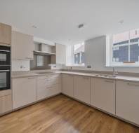 L-shaped kitchen with light-colored cabinets, wood flooring, a built-in double oven, and two windows looking out onto a dark-sided building.