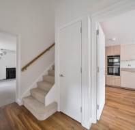 A hallway with carpeted stairs, a central white door, and doorways leading to a living room with a fireplace and a kitchen with light wood cabinetry.