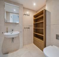 A bathroom with a wall-mounted sink, a mirror, two sets of coat hooks, a wooden shelving unit with two drawers, and a toilet.