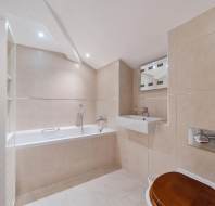 A modern bathroom with beige tiled walls, a white bathtub with a handheld shower head, a wall-mounted sink, and a toilet with a dark wood seat.