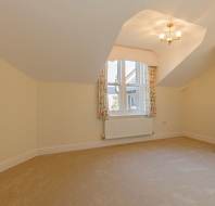 An empty room with cream walls, beige carpeting, a window with floral curtains, and a ceiling light fixture.