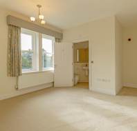 Empty room with cream carpet and walls, two windows with patterned curtains, and an open door leading to a bathroom with a toilet and sink.