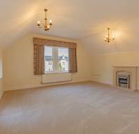 An empty room with cream walls, carpeted flooring, two chandeliers, a central window with floral curtains, and a fireplace.