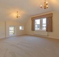 An empty room with cream walls, carpeted flooring, two chandeliers, and windows overlooking neighboring buildings.