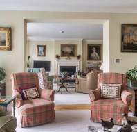 A living room featuring two matching plaid armchairs with personalized pillows, a fireplace in the background, and multiple paintings on the walls.