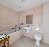 A tiled bathroom with a bathtub, pedestal sink, wall-mounted mirror, toilet, and a wicker basket filled with towels.
