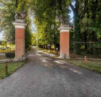 An asphalt driveway leads through brick gate pillars topped with stone lion statues, flanked by black metal fencing and a tree-lined path.
