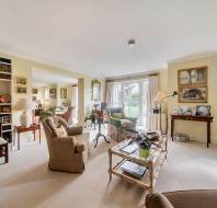 A bright, furnished living room featuring a floor-to-ceiling bookshelf, multiple armchairs, a glass-topped coffee table, and French doors leading to a garden.