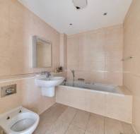Bathroom with beige tiled walls and floors, featuring a wall-hung sink, a bidet, a bathtub with a handheld shower head, and a chrome towel radiator.
