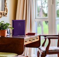 A wooden desk in front of a window holding a potted plant, a purple folder with the Audley Villages logo, and a stack of small books, with a wooden chair placed to the side.