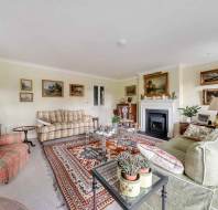 A living room featuring a plaid sofa and armchair, a green sofa, a large patterned area rug, a glass coffee table, and a fireplace with paintings displayed on the walls.