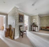 A hallway with light green floral wallpaper and beige carpet, featuring a wooden chair, a console table, and doorways leading to other rooms.