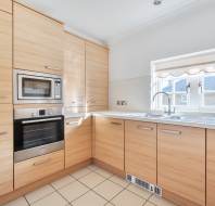 L-shaped kitchen with light wood cabinets, stainless steel appliances including a built-in microwave and oven, a double sink under a window, and tiled flooring.