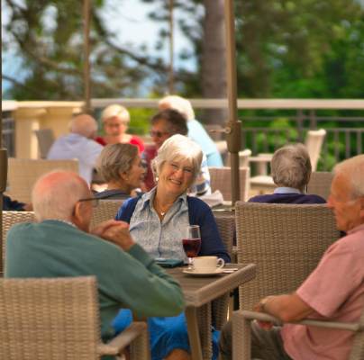 A group of older adults sit at outdoor tables on a patio, talking and drinking, with trees and a railing visible in the background.