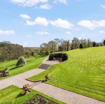 A landscaped garden with manicured lawns, stone pathways, and wooden benches surrounded by trees and hills on a clear day with a blue sky and white clouds.