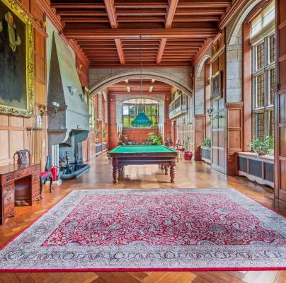A spacious, wood-paneled room with a billiards table, ornate rug, large windows, a fireplace, and elegant furniture, including a desk and chairs, under a coffered ceiling.