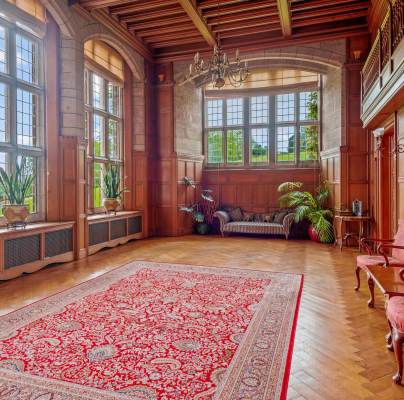 A large room features wood-paneled walls, tall arched windows, a red patterned rug, two upholstered chairs, and several potted plants. A chandelier hangs from the high ceiling.