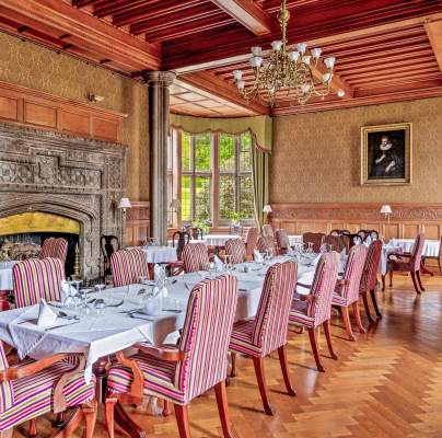 Elegant dining room with striped upholstered chairs, neatly set tables with white tablecloths, a large ornate fireplace, wooden paneling, and a chandelier. A portrait hangs on the wall near windows.