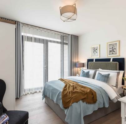 A neatly arranged bedroom with a double bed, light blue bedding, two bedside lamps, framed artworks, and a chair with a patterned blanket. The room has floor-to-ceiling windows with sheer curtains.