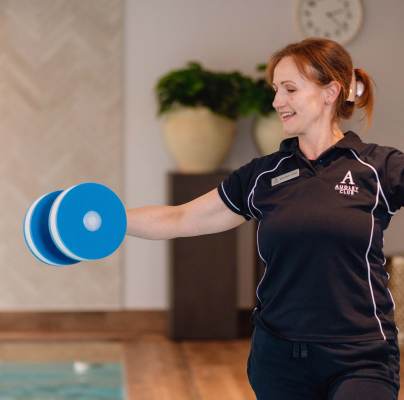 A woman in a uniform holds blue foam dumbbells with arms extended, standing indoors near a pool.