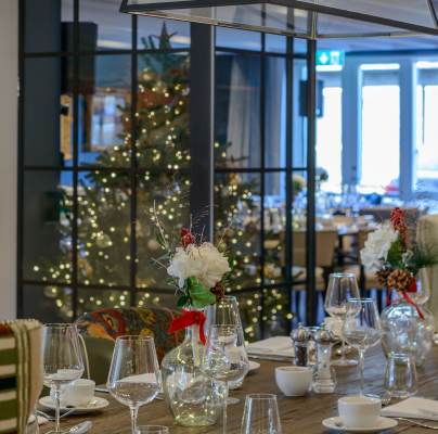 A festive dining table set with glassware and floral centerpieces. A decorated Christmas tree is visible in the background.