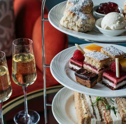 A three-tiered afternoon tea set with sandwiches, various cakes, scones with cream and jam. Two glasses of champagne are on the side.