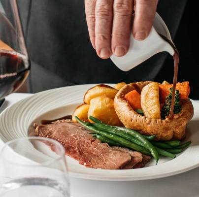 A hand pours sauce over a plate with roast beef, green beans, Yorkshire pudding, potatoes, and vegetables. A glass of red wine is on the table.
